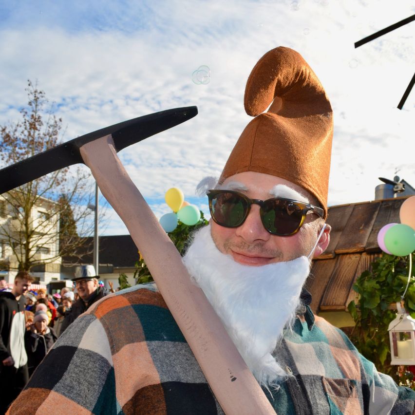 Ein Mann, verkleidet als Zwerg mit weißem Bart und Spitzhacke, lächelt in die Kamera. Er trägt einen braunen, spitzen Hut und eine Sonnenbrille. Hinter ihm befindet sich eine festliche Umgebung mit Luftballons und einer Menschenmenge.