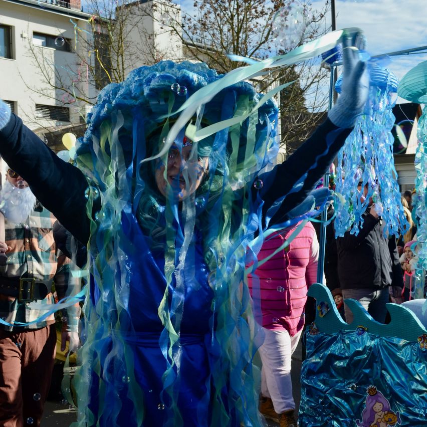 Eine Person in einem Quallenkostüm winkt mit blauen Bändern bei einem Straßenfest, umgeben von anderen Teilnehmern.