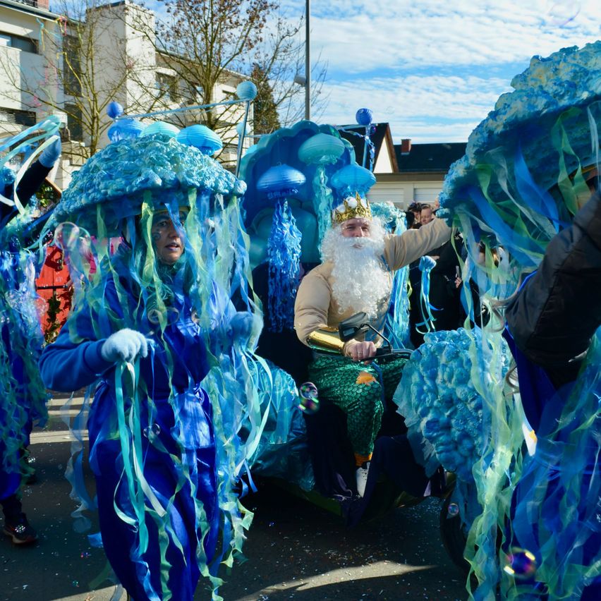 Eine Gruppe von Menschen in blauen Kostümen mit quallenartigen Kopfbedeckungen, einer von ihnen als Meereskönig verkleidet, nimmt an einem Umzug teil.