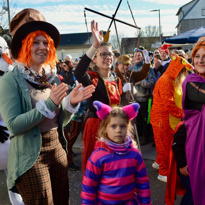 Eine Menschenmenge in Kostümen ist im Freien versammelt. Eine Frau in einer Mad Hatter-Kostüm lächelt und klatscht. Ein Mädchen in einem gestreiften Cat in the Hat-Kostüm steht vor ihr. Hinter ihnen tragen andere verschiedene Kostüme, und einige Gebäude sind sichtbar.