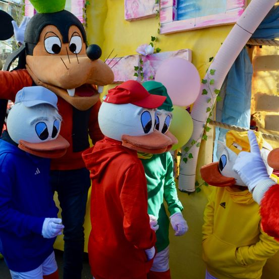 Eine Gruppe von Maskottchen, einschließlich Micky Maus, gekleidet in Kostümen und nebeneinander stehend. Hinter ihnen ist eine farbenfrohe Wand mit Luftballons und dekorativen Elementen.
