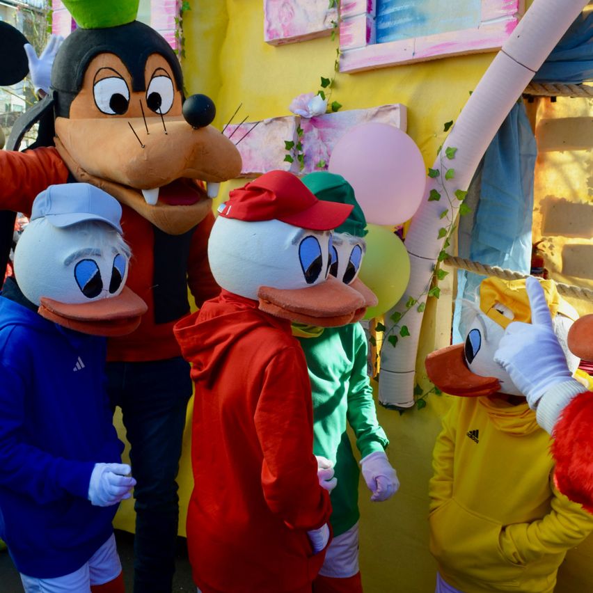 Eine Gruppe von Maskottchen, einschließlich Micky Maus, gekleidet in Kostümen und nebeneinander stehend. Hinter ihnen ist eine farbenfrohe Wand mit Luftballons und dekorativen Elementen.