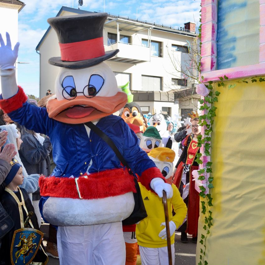 Ein Maskottchen winkt bei einer Parade mit anderen Maskottchen, umgeben von Zuschauern in Kostümen.