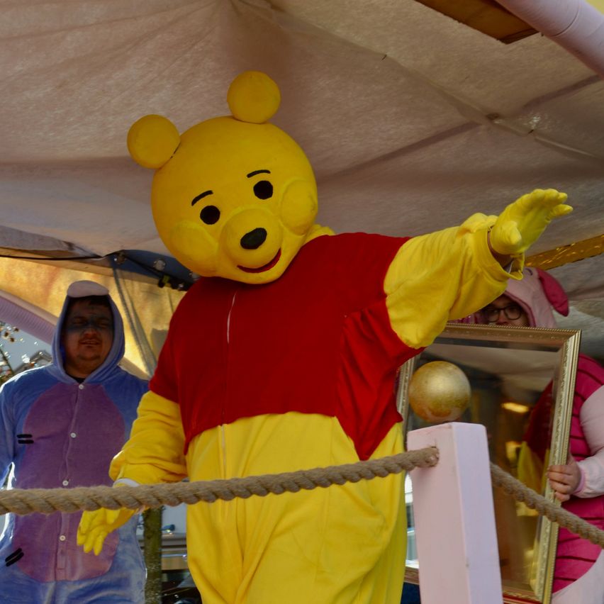 Eine Person in einem Winnie-Pooh-Kostüm winkt in die Kamera. Dahinter steht eine weitere Person in einer blauen Kapuzenjacke und eine dritte in einer rosa Kleidung. Ein Spiegel reflektiert einen goldenen Ball.