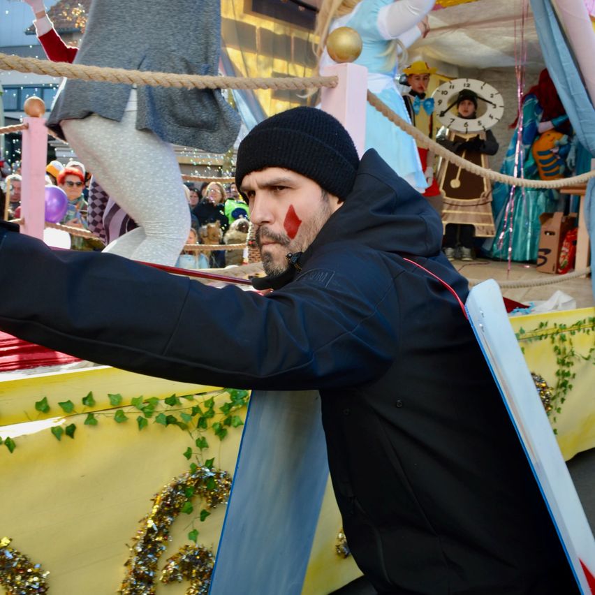 Ein Mann in einem schwarzen Mantel und Mütze steht auf einem Karnevalswagen, hält einen Schild und hat vor sich ein Seil, während Menschen in Kostümen hinter ihm stehen.
