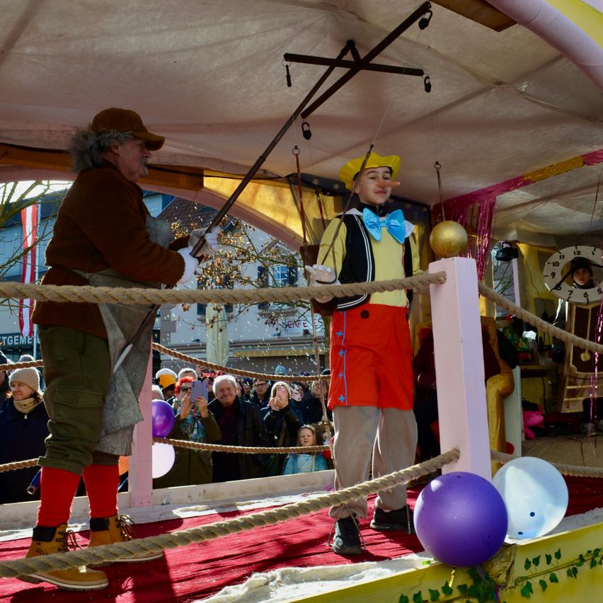 Zwei Darsteller auf einer Bühne, einer hält eine Marionette und der andere ist als Clown verkleidet, stehen vor einer Menge auf einem Karnevalswagen. Die Bühne ist mit Seilen, Luftballons und einem roten Teppich dekoriert. Hinter ihnen sind eine Uhr und verschiedene Requisiten zu sehen.