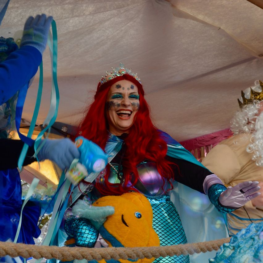 Eine Frau in einem Meerjungfrauen-Kostüm lächelt und hält einen gelben Fisch. Hinter ihr hält eine Person in einem blauen Handschuh eine blaue Schleife. Eine andere Person mit einer weißen Perücke und einer Krone steht hinter ihr.