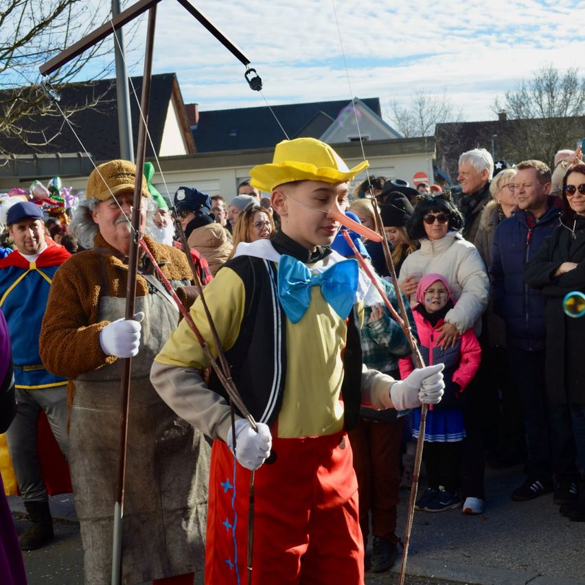 Eine Gruppe von Menschen in verschiedenen Kostümen steht im Freien. Eine Person in einem gelben Hut und roten Hosen steht in der Mitte. Dahinter stehen einige Menschen und beobachten.