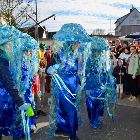 Eine Gruppe von Menschen, die blaue Kostüme tragen, die Quallen ähneln, gehen auf der Straße, während eine Menge Menschen vom Bürgersteig zusieht.