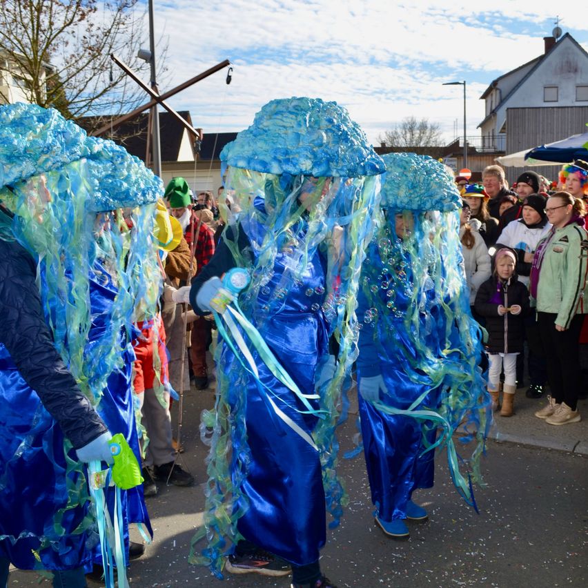 Eine Gruppe von Menschen, die blaue Kostüme tragen, die Quallen ähneln, gehen auf der Straße, während eine Menge Menschen vom Bürgersteig zusieht.