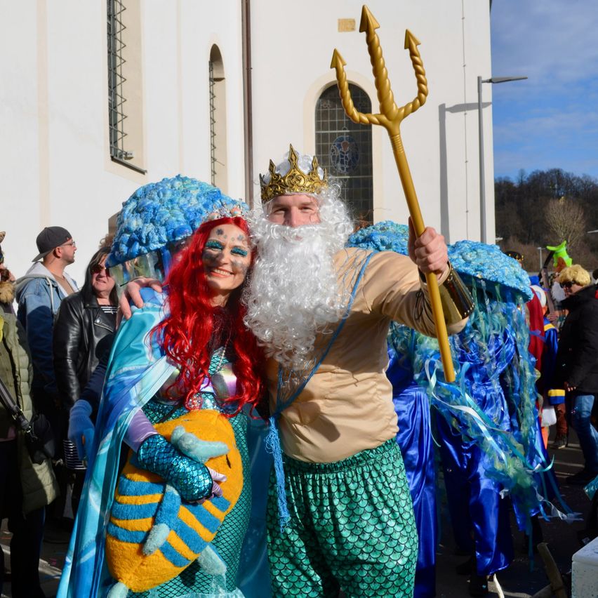 Ein Mann und eine Frau, gekleidet als König und Königin des Meeres, stehen zusammen mit einer Menschenmenge in einer Festumgebung.