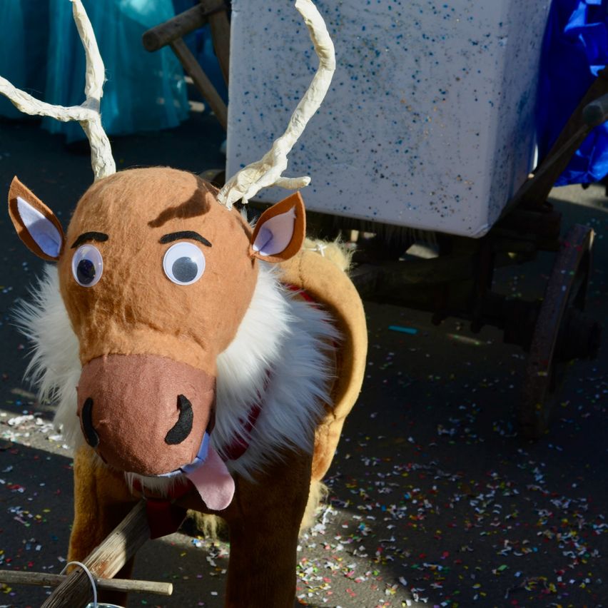 Ein stofftier-rentier steht auf einer straße, mit buntem konfetti darum verstreut, und ein hölzerner wagen mit einer weißen box dahinter.