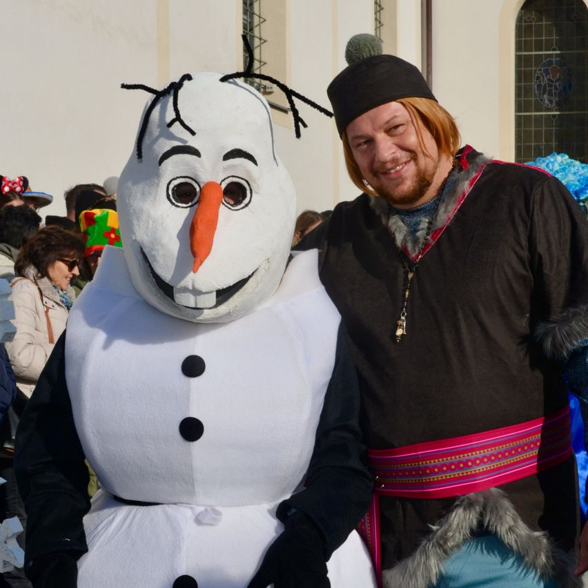 Eine Person in einer Olaf-Kostüme aus Frozen steht neben einem Mann in traditioneller skandinavischer Kleidung. Sie lächeln und posieren für ein Foto, mit einer Menge Menschen im Hintergrund.