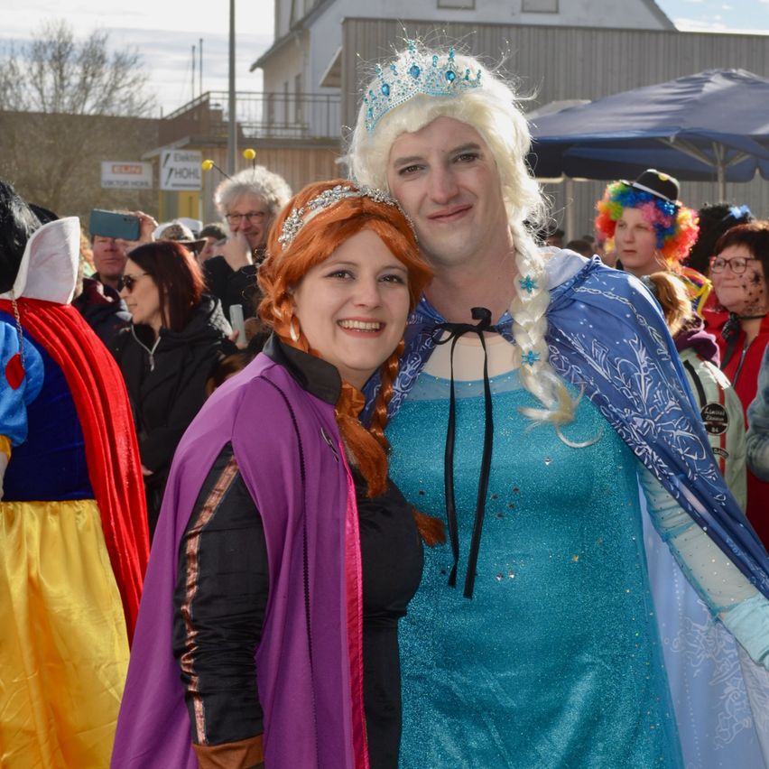 Ein Mann und eine Frau in Disney-Prinzessinnenkostümen stehen lächelnd in einer Menschenmenge. Dahinter befinden sich Menschen in verschiedenen Kostümen und ein Gebäude mit Balkon.