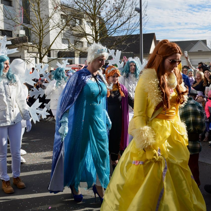 Eine Gruppe von Menschen in Kostümen, wahrscheinlich für einen Umzug, geht auf der Straße. Einige sind als Disney-Charaktere verkleidet. Ein Gebäude mit Balkonen und Bäumen im Hintergrund.