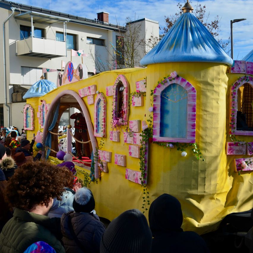 Ein gelber Umzugswagen mit einer blauen Turmspitze und rosa Akzenten ist bei einem Umzug. Menschen schauen zu, einige tragen Hüte. Eine Figur steht drinnen, mit Ballons und Girlanden. Gebäude und Bäume sind im Hintergrund.