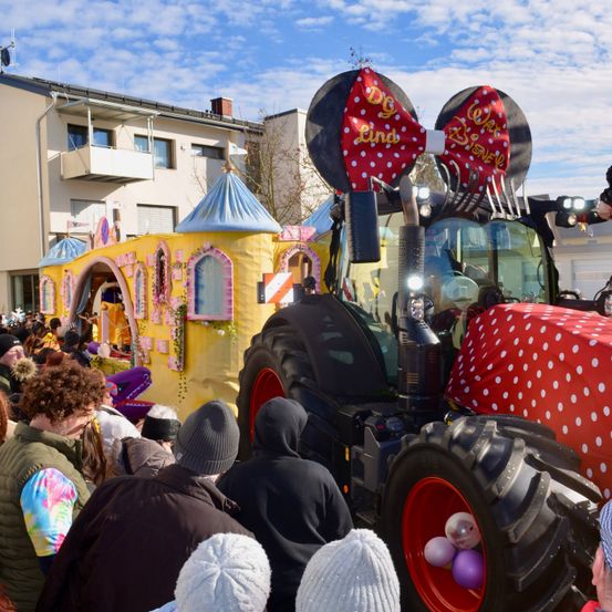 Eine große Menschenmenge beobachtet einen geschmückten Traktor mit Minnie-Mouse-Ohren bei einer Veranstaltung im Freien, mit Gebäuden und blauem Himmel im Hintergrund.