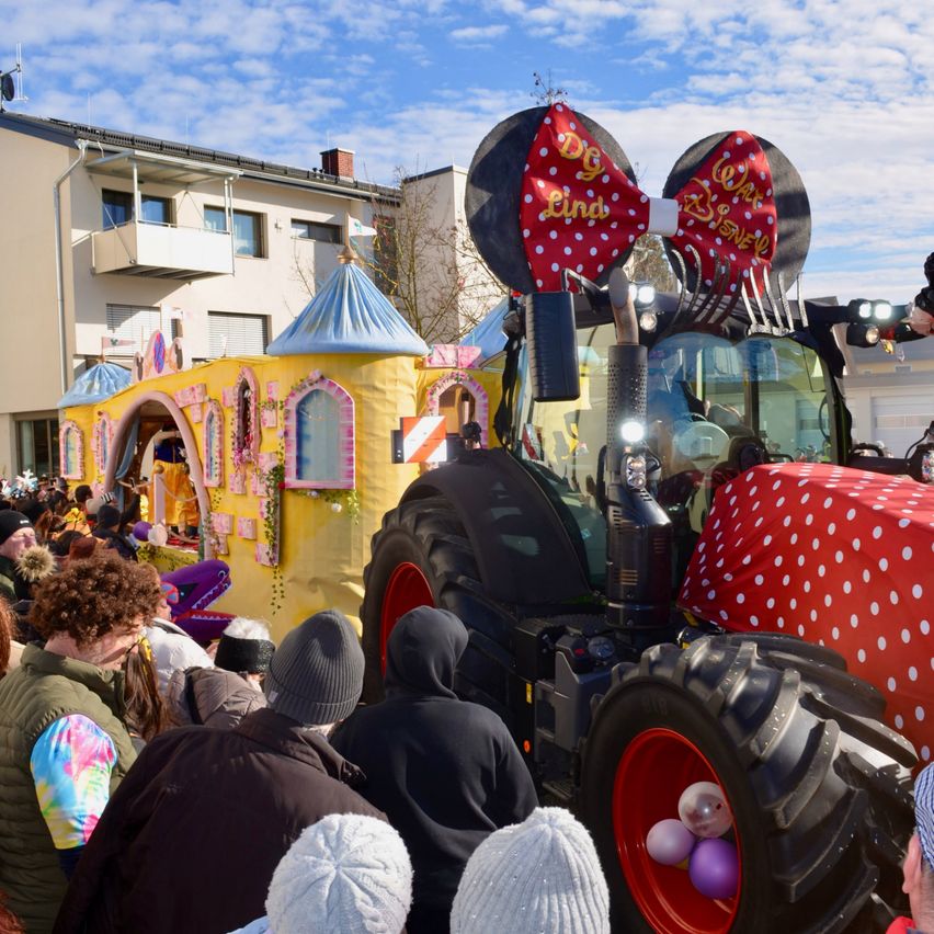 Eine große Menschenmenge beobachtet einen geschmückten Traktor mit Minnie-Mouse-Ohren bei einer Veranstaltung im Freien, mit Gebäuden und blauem Himmel im Hintergrund.