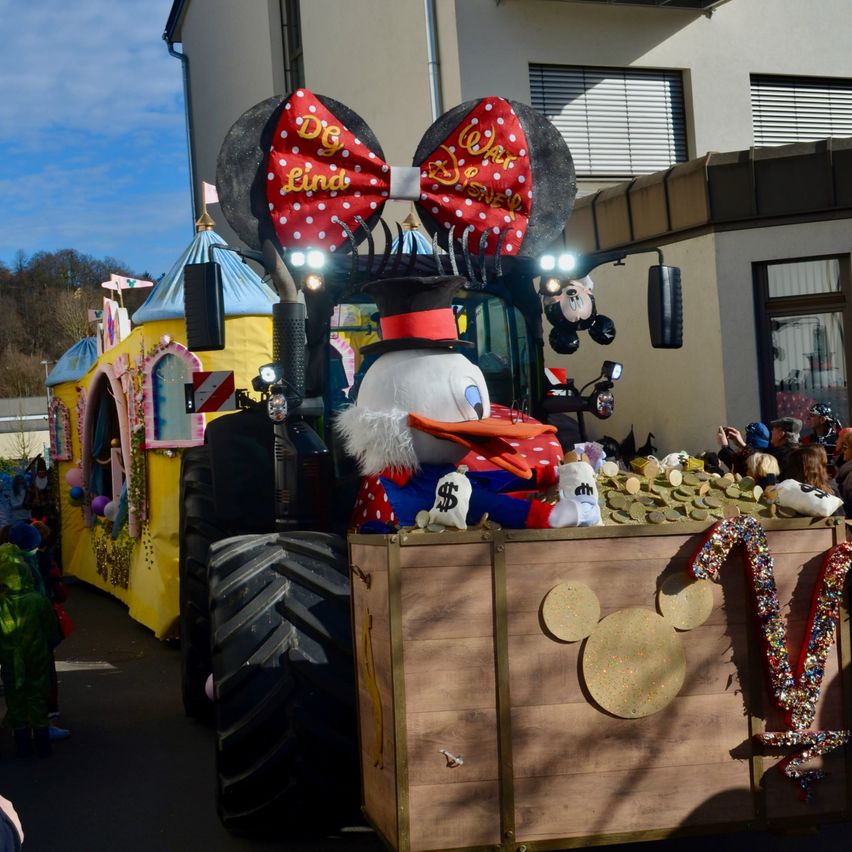 Ein Umzugswagen mit einem Micky-Maus-Traktor, in dem Goofy in einer Schatztruhe sitzt, verziert mit Münzen und Micky-Maus-Ohren, vor einem gelben Gebäude mit Zuschauern.