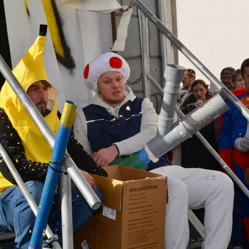 Zwei Männer in Kostümen sitzen auf einer Treppe. Einer trägt eine Bananen-Verkleidung und der andere eine rot-weiße Kappe. Ein Karton liegt auf der Treppe. Dahinter beobachtet eine Menge.
