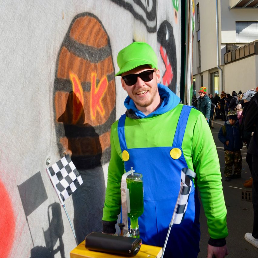Ein Mann in einem grünen Mario-Kostüm steht neben einer Graffiti-Wand mit einem Fass, hält ein grünes Getränk und eine gemusterte Flagge.