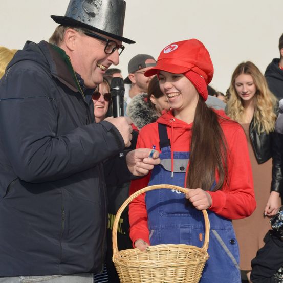 Ein Mann in einem schwarzen Hut reicht einer lächelnden Frau, die eine rote Mütze trägt, einen Korb. Dahinter beobachtet eine Menschenmenge. Die Frau hält einen Stift und der Korb ist braun.