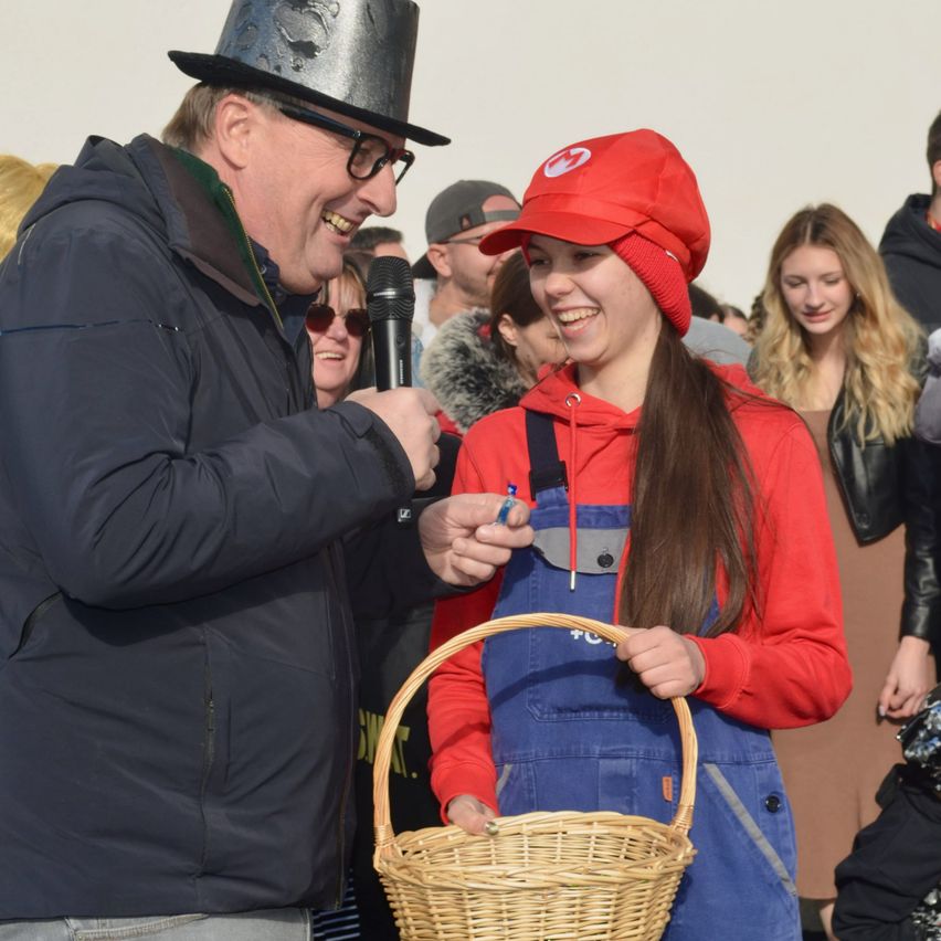 Ein Mann in einem schwarzen Hut reicht einer lächelnden Frau, die eine rote Mütze trägt, einen Korb. Dahinter beobachtet eine Menschenmenge. Die Frau hält einen Stift und der Korb ist braun.