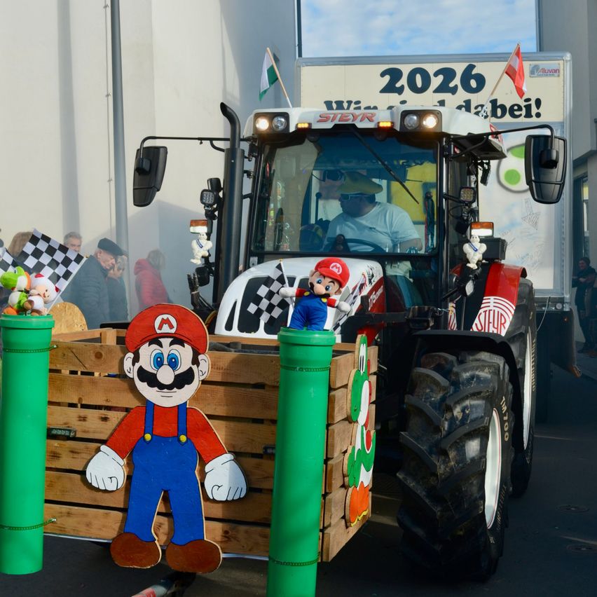 Ein Traktor mit einem Mario-thematischen Anhänger ist geparkt. Ein Mann fährt ihn. Flaggen sind an der Front montiert. Hinter dem Traktor zeigt ein LKW eine bunte Werbung an.