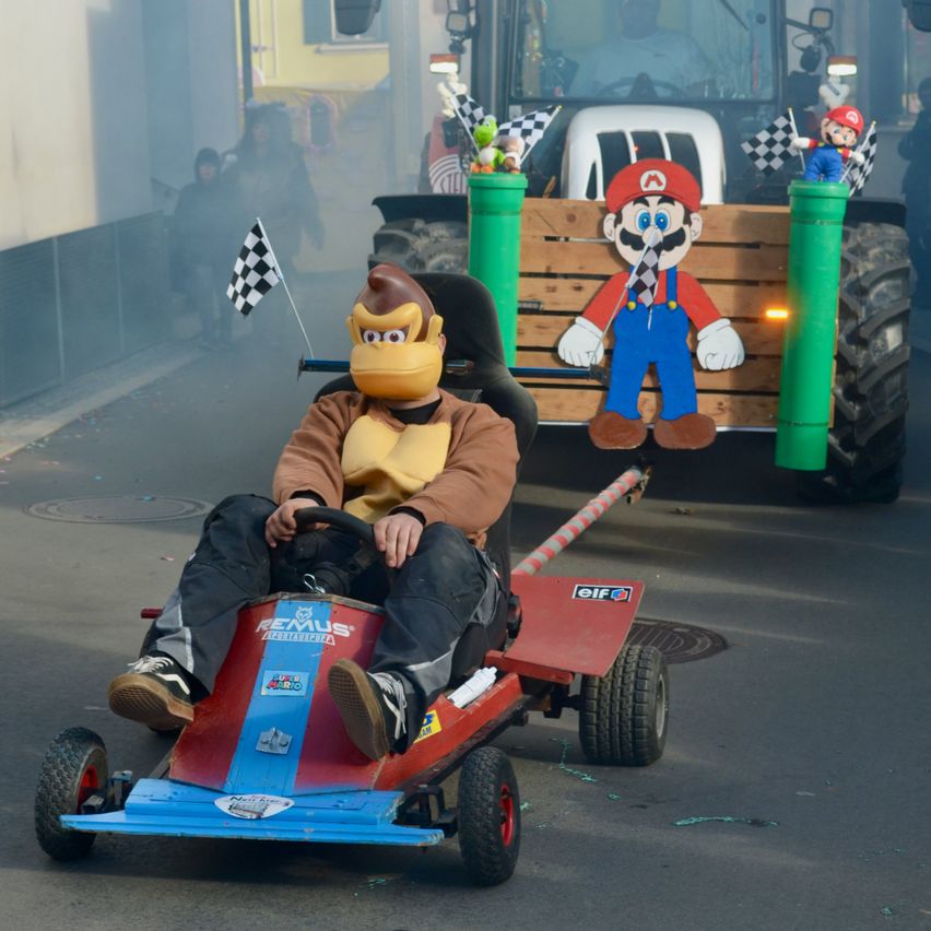 Eine Person mit einer Donkey-Kong-Maske fährt einen Go-Kart, hinter dem ein Traktor steht. Auf dem Traktor steht eine Mario-Figur, mit Rauch und Fahnen im Hintergrund.