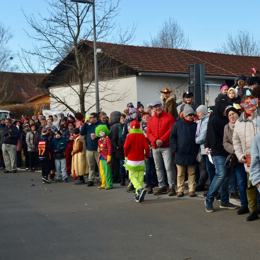 Viele Menschen sind in einer festlichen Umgebung versammelt, einige in Kostümen. Ein Gebäude mit einem braunen Dach ist im Hintergrund zu sehen.