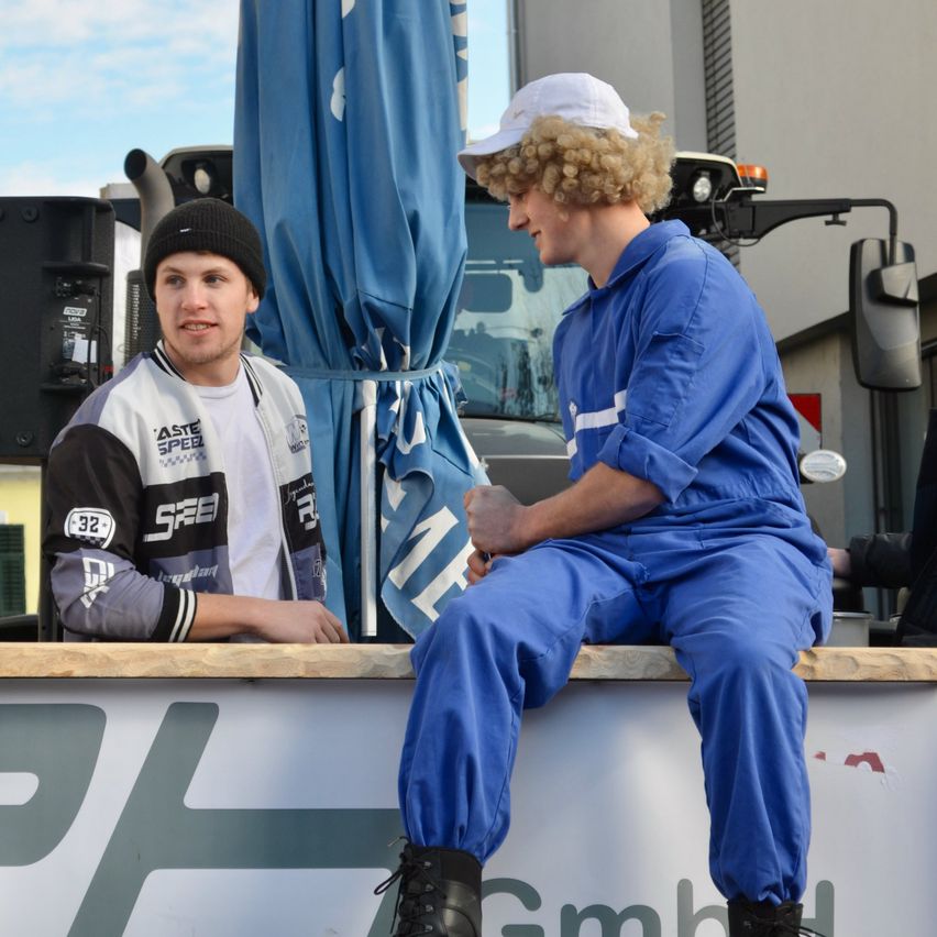 Zwei Männer in blauen Arbeitsuniformen sitzen auf einer hölzernen Kante in der Nähe eines Fahrzeugs. Einer trägt eine weiße Kappe und hat lockiges Haar, der andere trägt eine schwarze Mütze. Hinter ihnen ist ein blauer Banner.