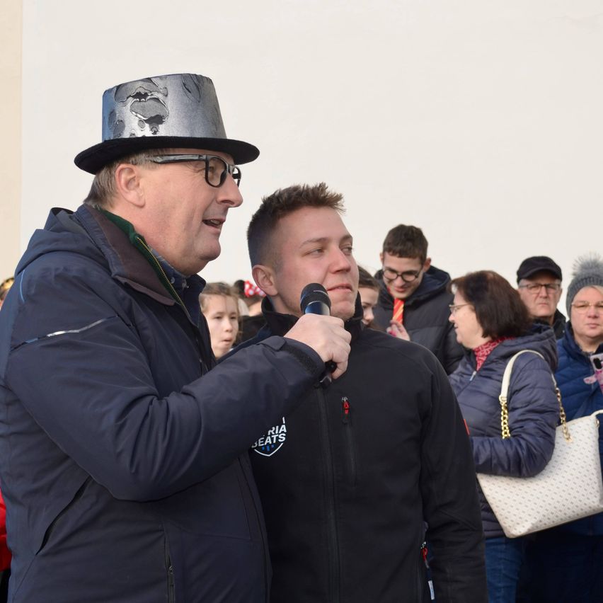 Ein Mann mit einem silbernen Zylinderhut hält ein Mikrofon und spricht mit einem Mann in einer blauen Jacke in einer Menschenmenge.