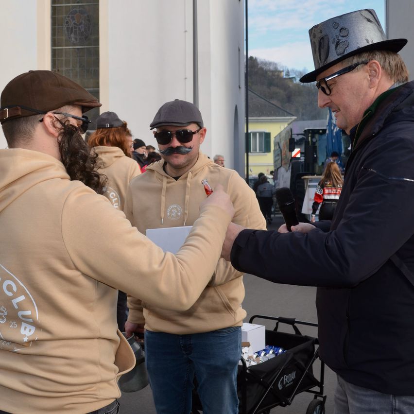 Zwei Männer mit Hüten und Sonnenbrillen unterhalten sich, einer hält ein Mikrofon. Sie stehen vor einem Gebäude mit einem Trolley in der Nähe.