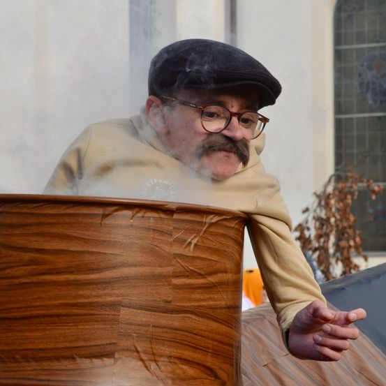 Ein älterer Mann mit Schnurrbart und Brille steht neben einem Holzfass und bläst Rauch aus. Er trägt einen schwarzen Filzhut und eine beigefarbene Kapuzenjacke.