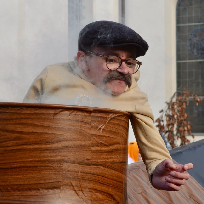 Ein älterer Mann mit Schnurrbart und Brille steht neben einem Holzfass und bläst Rauch aus. Er trägt einen schwarzen Filzhut und eine beigefarbene Kapuzenjacke.