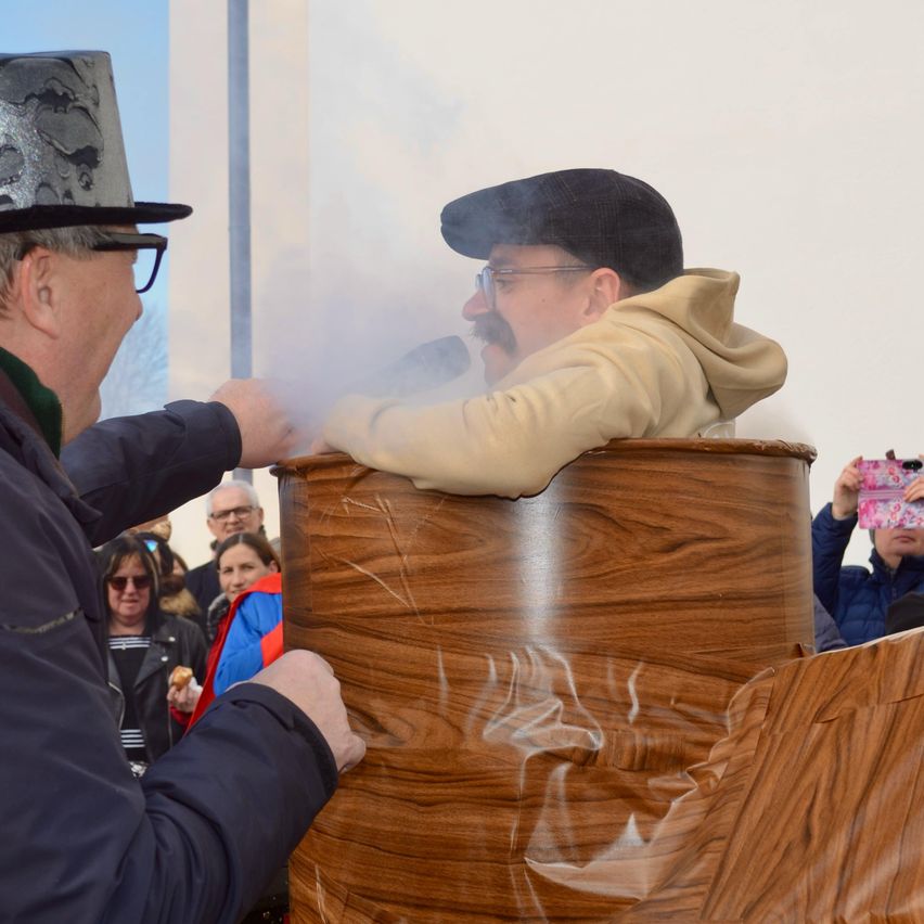 Ein Mann mit Hut raucht eine Pfeife in einem Fass, während ein anderer das Fass hält. Im Hintergrund sehen Menschen zu und fotografieren.