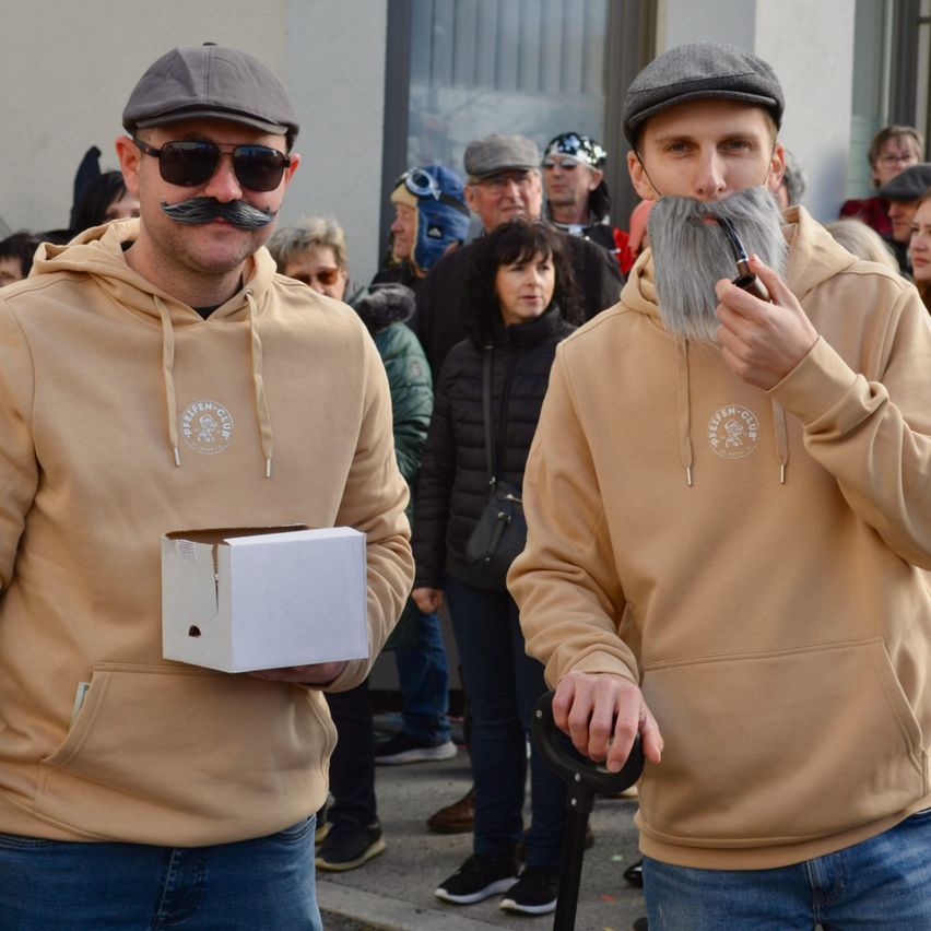 Zwei Männer mit falschen Schnurrbärten und Bärten stehen vor einer Menge. Einer hält eine Box und trägt einen Hut, der andere raucht eine Pfeife.