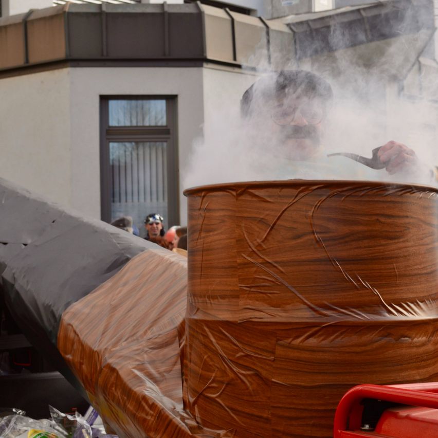 Ein Mann mit Schnurrbart sitzt auf einem hölzernen Fass, umgeben von Rauch, vor einem Gebäude mit Fenster und einer Gruppe von Menschen.