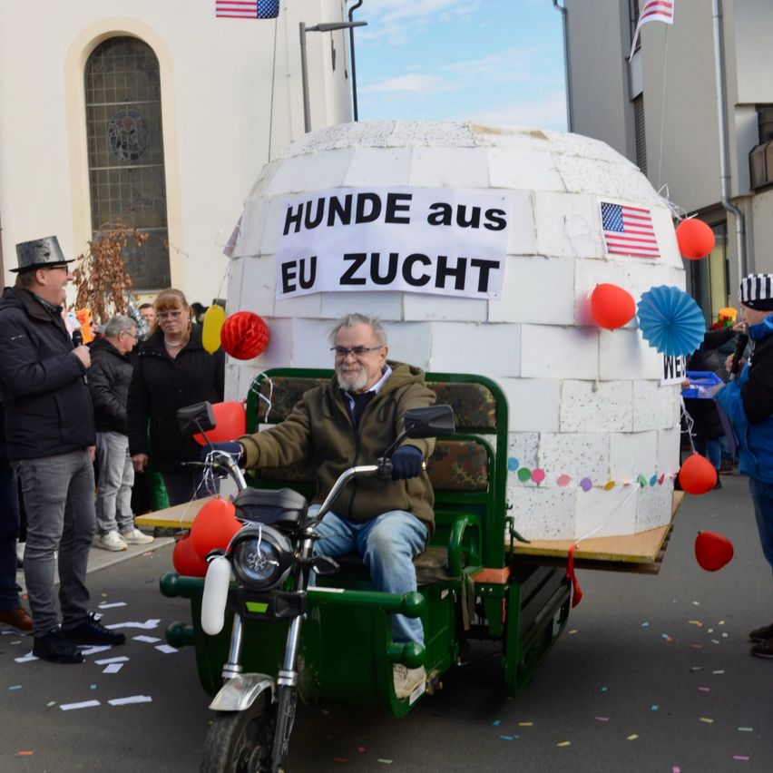 Ein Mann fährt auf einem motorisierten Wagen mit einem Anhänger in Form eines Hauses. Dahinter hängt ein Banner mit der Aufschrift 'HUNDE aus EU ZUCHT'. Es gibt Ballons und Papierdekorationen. Zuschauer beobachten.