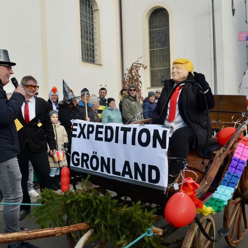 Eine Gruppe von Menschen in Kostümen versammelt sich um eine Kutsche mit einem Schild, auf dem Expedition Grönland steht. Eine Person in einer Trump-Maske sitzt in der Kutsche.