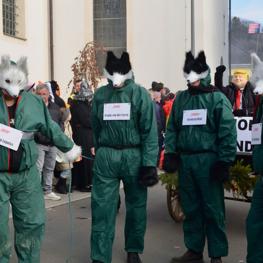 Mehrere Personen in grünen Anzügen und Tiermasken stehen in einer Reihe. Die Person auf der linken Seite trägt eine Fuchsmaske. Die Person in der Mitte hat ein Schild mit der Aufschrift 'Ersula von der Leyen'. Die Person auf der rechten Seite hat ein Schild mit der Aufschrift 'Freddie'. Eine Menschenmenge steht hinter ihnen.