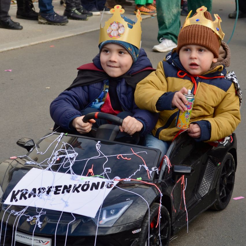 Zwei kleine Jungen sitzen in einem dekorierten schwarzen Auto und tragen Kronen. Einer hält eine Dose Limonade. Ein Banner auf dem Auto steht 'KRAPFENKONIG'. Hinter ihnen laufen Leute auf einer Straße.