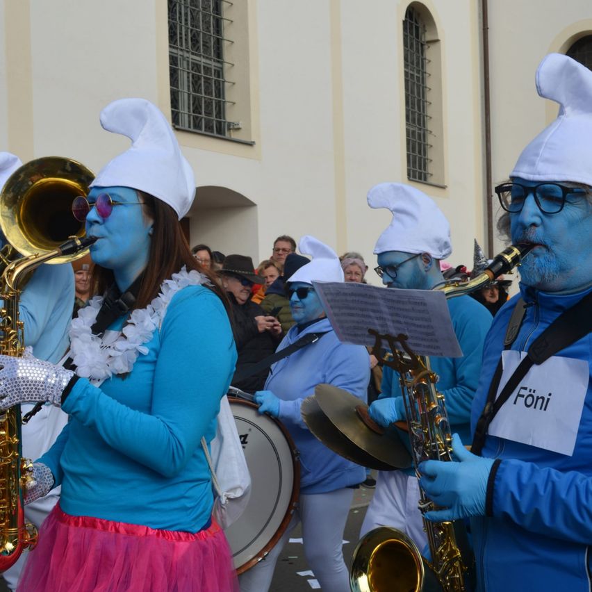 Eine Gruppe von Menschen, die in blauer Gesichtsbemalung und weißen Hüten gekleidet sind, spielt auf Musikinstrumenten, einschließlich Saxophonen und einer Trommel, in einer Außenumgebung.