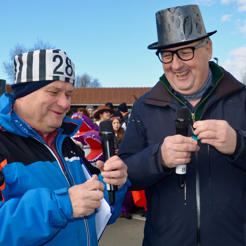 Zwei Männer in Winterkleidung, einer mit einem gestreiften Hut und der andere mit einem Hut und einem Mikrofon, stehen nebeneinander und lächeln.