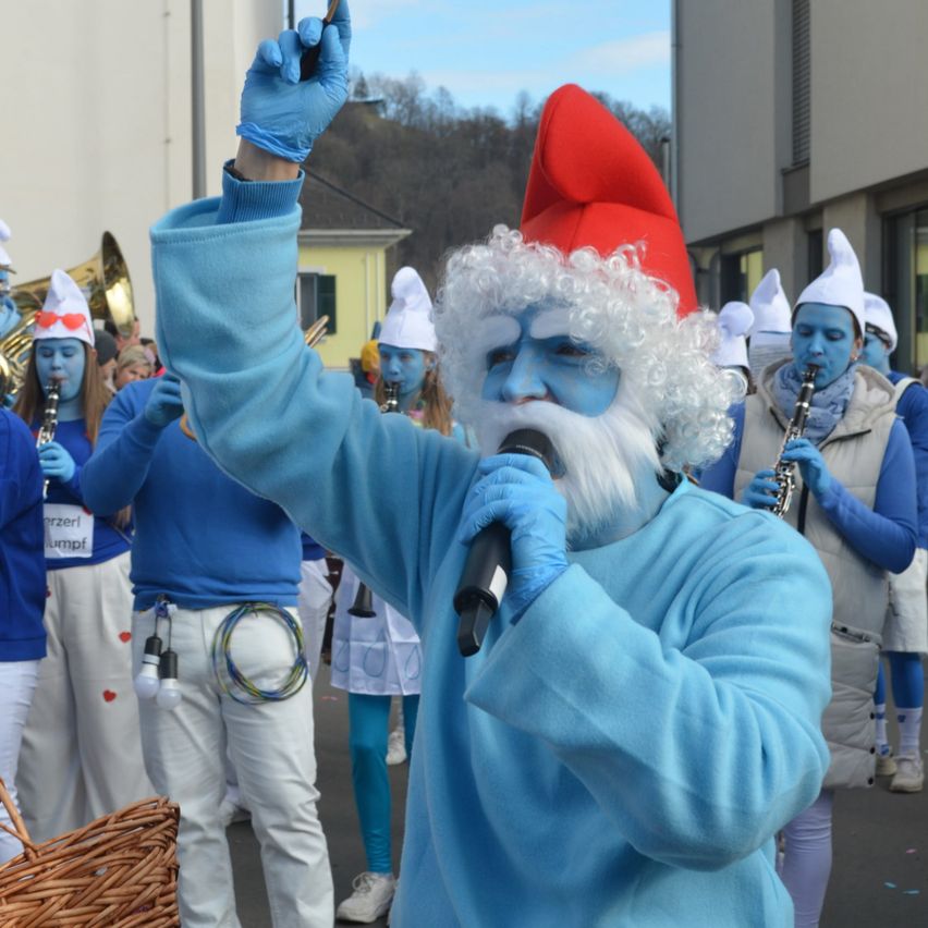 Eine Person in einem blauen Kostüm und weißem Bart führt eine Gruppe von ähnlich gekleideten Personen bei einem Umzug an. Sie halten Musikinstrumente und eine Person hält ein Mikrofon.