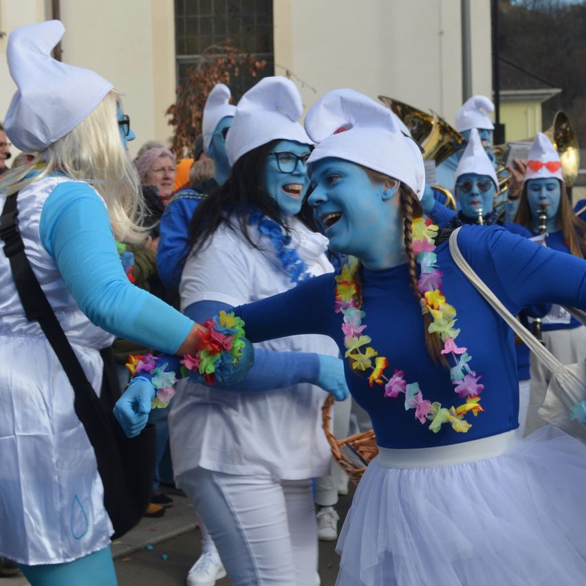 Mehrere Menschen in blauer Gesichtsbemalung und Kostümen tanzen draußen zusammen. Sie tragen weiße Hüte, Blumenketten und blaue Handschuhe. Eine Menge Zuschauer steht hinter ihnen.