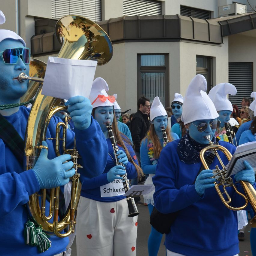 Eine Gruppe von Menschen mit blauer Gesichtsbemalung, Handschuhen und Mützen spielt Instrumente bei einem Outdoor-Umzug. Ein Mann spielt Tuba, während andere Trompeten und Klarinetten spielen.