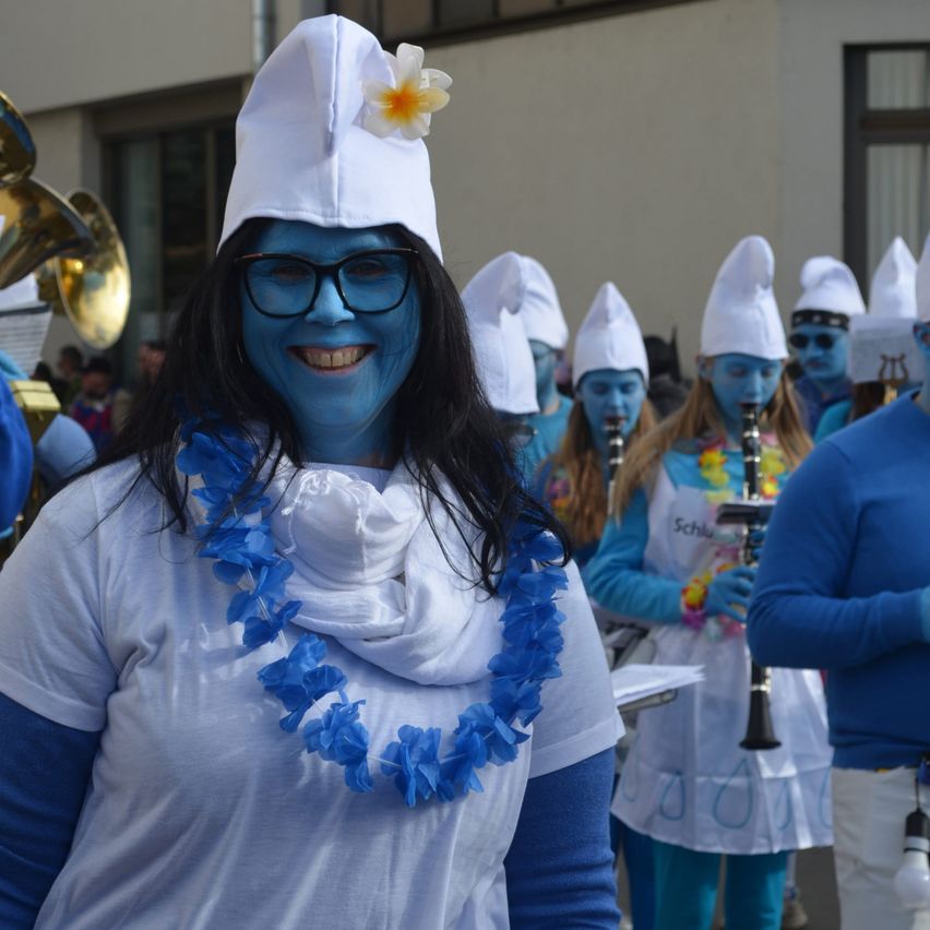 Eine Frau mit einem weißen Hut, einer Blume und blauer Gesichtsbemalung lächelt für die Kamera. Hinter ihr spielt eine Gruppe von Menschen in blauen Kostümen und weißen Hüten Musikinstrumente.