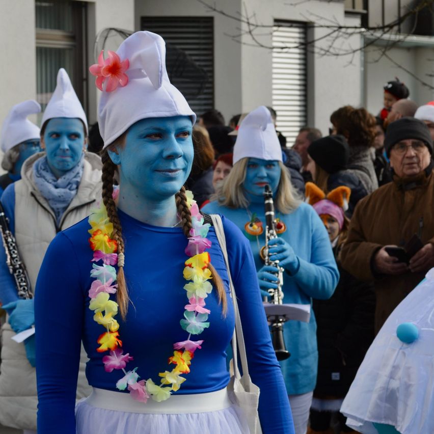 Mehrere Personen tragen blaue Körperbemalung und Kostüme. Eine Frau vorne trägt ein blaues Shirt, einen weißen Rock und eine Blumenkette. Dahinter hält eine Person eine Klarinette.