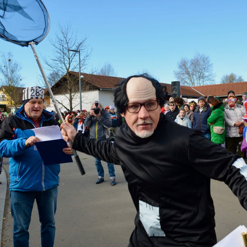 Ein Mann mit schwarzer Perücke und Brille hält eine Netz in einer Parade, ein anderer Mann gibt ihm ein Papier, eine Menge beobachtet.